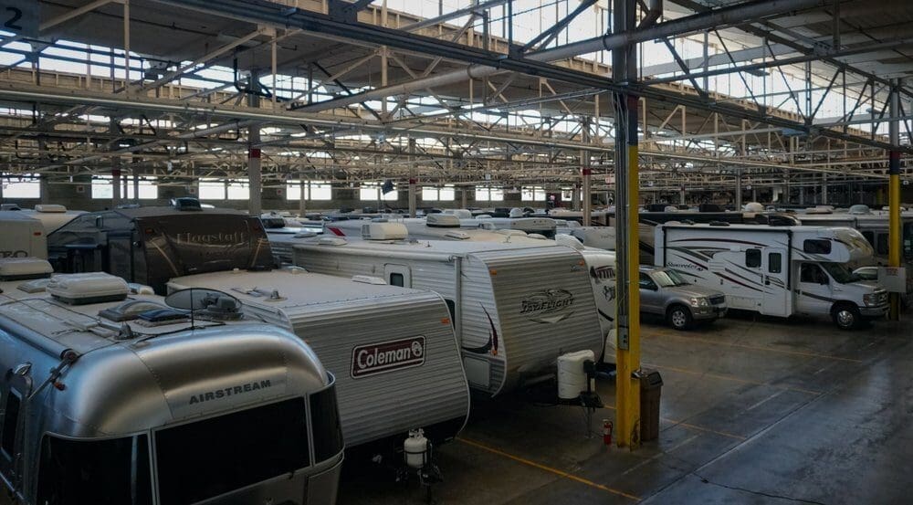 A large indoor storage facility with rows of parked RVs, including brands like Airstream and Coleman. High ceilings and industrial lighting create a spacious atmosphere.
