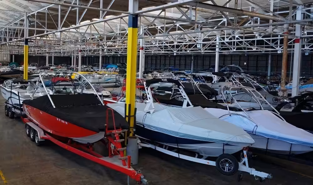 A spacious warehouse filled with numerous covered boats organized in rows. The boats are on trailers, ready for storage or transport, under a steel-beam roof.