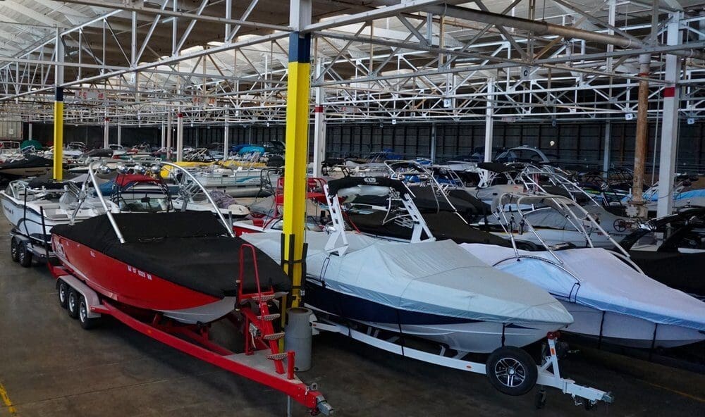 A spacious warehouse filled with numerous covered boats organized in rows. The boats are on trailers, ready for storage or transport, under a steel-beam roof.