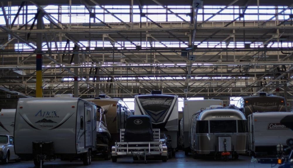 Inside a vast warehouse, various RVs and trailers are parked side by side. The ceiling features a grid of exposed beams and skylights. The atmosphere is industrial.
