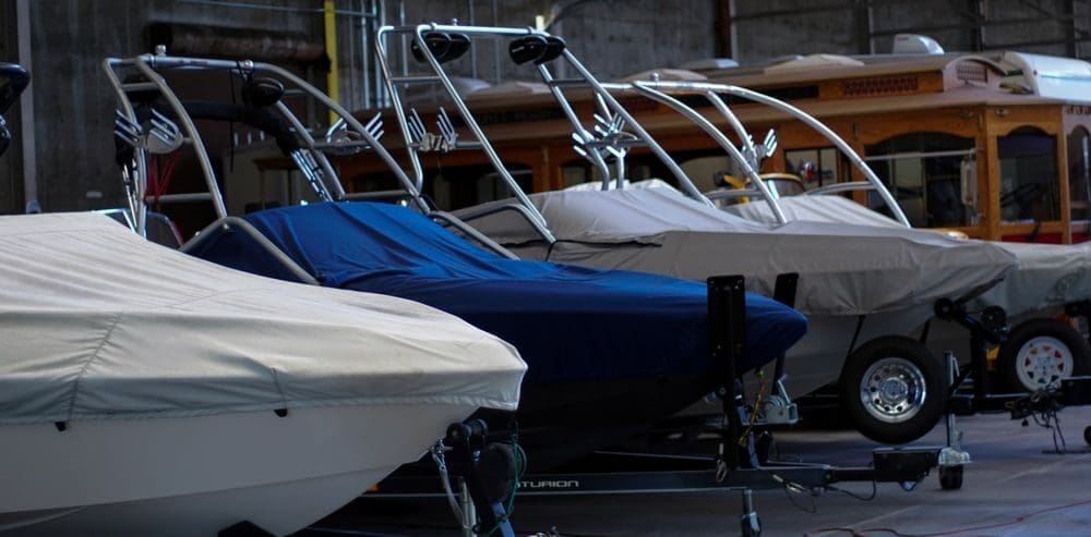 Several boats are parked indoors, covered in protective tarps. The dimly lit setting conveys a calm, organized storage environment.