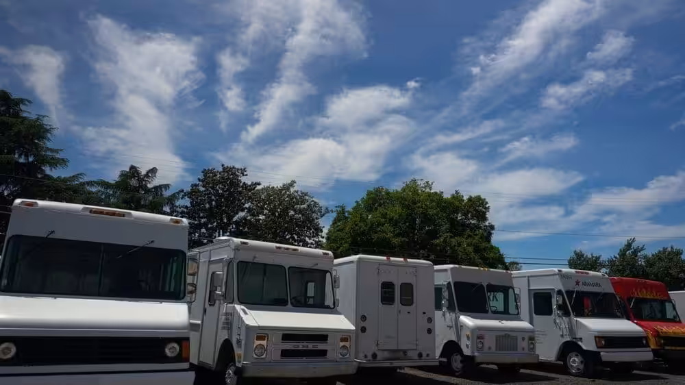 A line of six parked food trucks under a clear blue sky with wispy clouds. The trucks are white and red, surrounded by green trees, creating a serene scene.