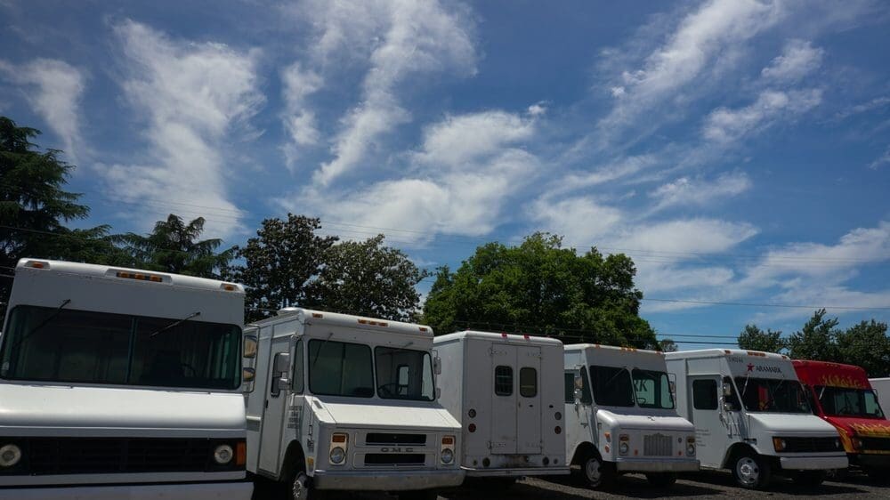 A line of six parked food trucks under a clear blue sky with wispy clouds. The trucks are white and red, surrounded by green trees, creating a serene scene.